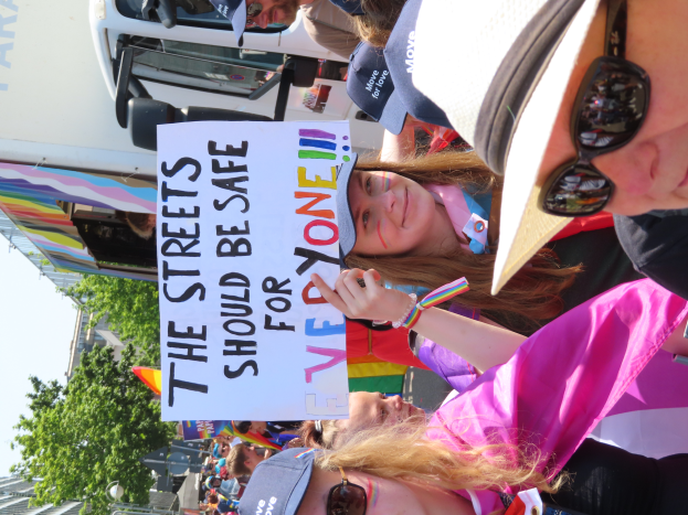 Eine Gruppe von Menschen steht vor einem Lastwagen und hält ein Schild mit der Aufschrift "Die Straßen sollten sicher sein". Einige tragen Mützen und Schutzbrillen, andere halten Schilder, vor einem Hintergrund aus Bäumen, Gebäuden und einem klaren blauen Himmel.