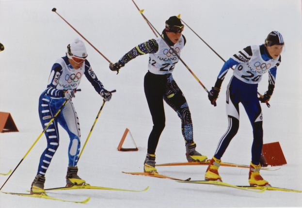 Drei Skifahrer sausen eine schneebedeckte Piste hinunter, jeder hält Ski-Stöcke und trägt bunte Ski-Anzüge und Helme, während die Sonne auf dem Schnee glitzert.