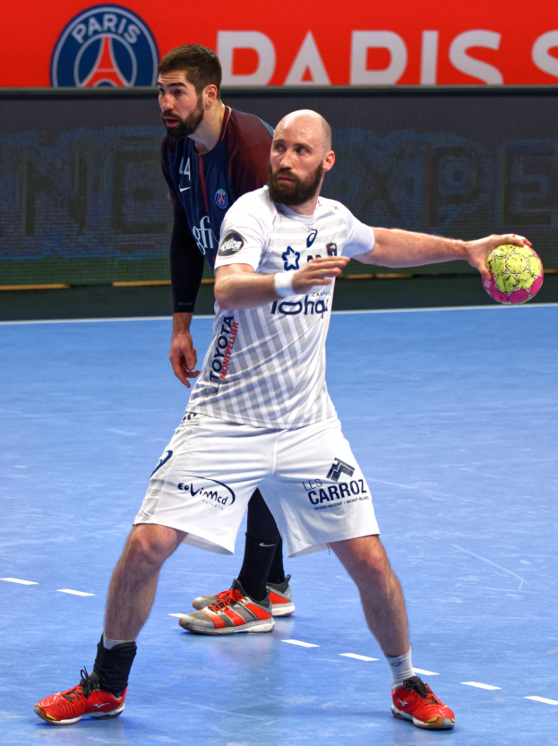 Zwei Männer beim Handballspielen auf einem Platz, einer hält den Ball, mit einem Hintergrundschild, auf dem "Paris Saint-Germain vs Paris Saint-Germain" steht.