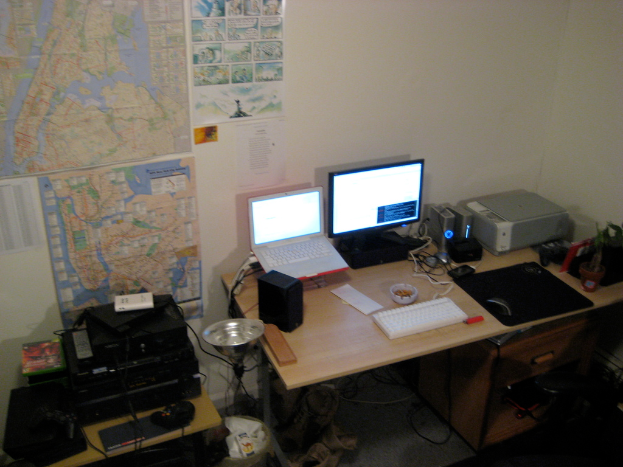 Ein Raum mit einem Tisch, auf dem ein Laptop, Lautsprecher, Monitor, Tastatur, Ball, Maus, Scanner und Papiere sowie ein weiterer Tisch mit Elektronikger├Ąten im Hintergrund stehen, mit Charts und Postern an der Wand dahinter.