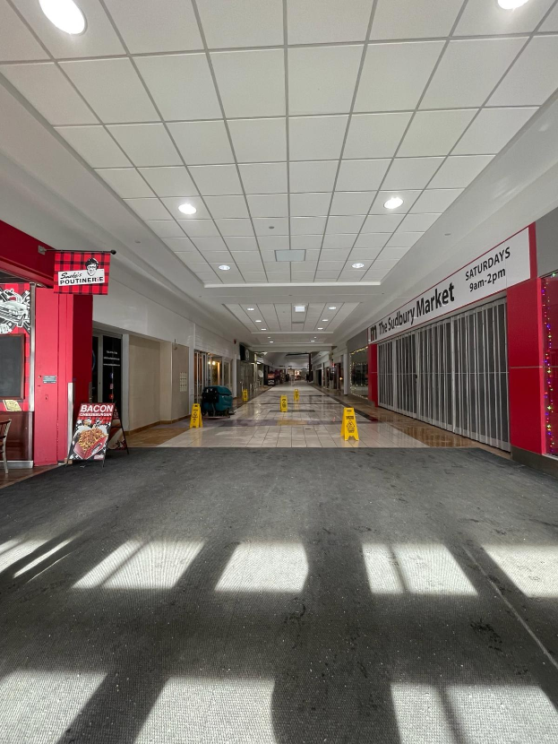 Innenansicht eines Einkaufszentrums mit Deckenbeleuchtung, einem Stuhl und einer Tafel mit der Aufschrift "Sydbury Market - Vorher und Nachher" links und verstreuten Gegenständen rechts.