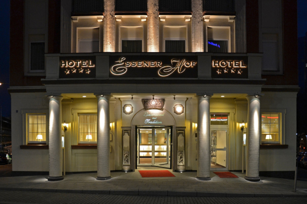Das Essener Hof Hotel in Berlin, Deutschland, mit einem Gebäude mit Fenstern, Türen, Säulen, Lampen und einem Namensschild, das entlang einer Straße mit sichtbaren Fahrzeugen und einem Himmel als Hintergrund zu sehen ist.