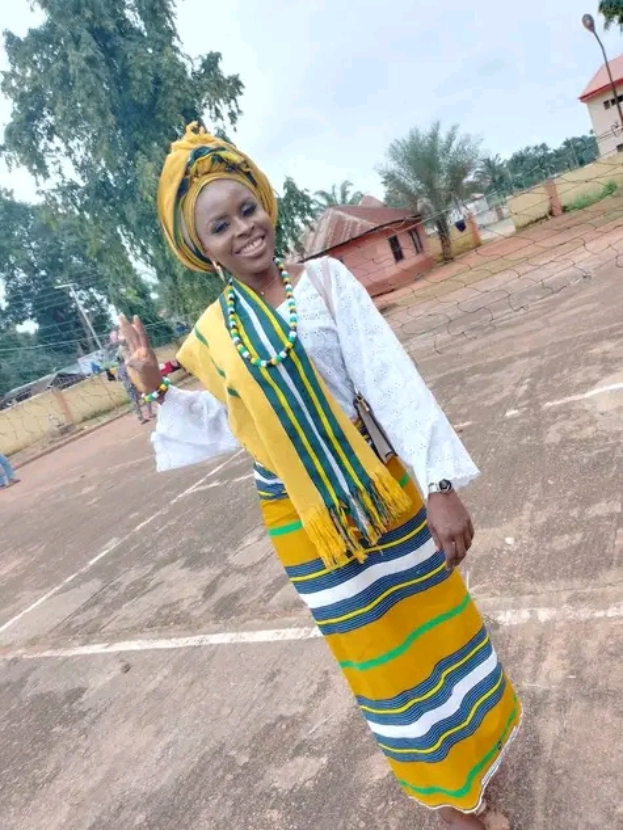 Eine Frau in einem gelben und blauen Kleid steht auf einem Basketballfeld, lächelt und posiert für ein Bild, mit Menschen, Häusern, Bäumen, Pfählen, Drähten und dem Himmel im Hintergrund.