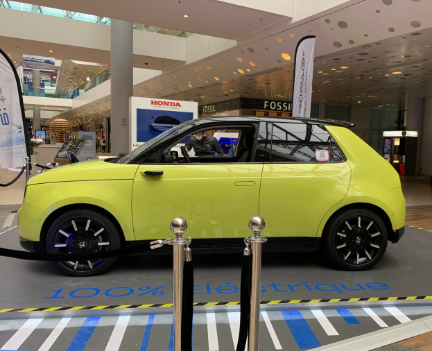 Gelber wasserstoffbetriebenes elektrisches Auto vor einem Gebäude mit Absperrpollern, Bannern, einer Person im Auto, einer Säule, stehenden Menschen, Schildern, einer Topfpflanze, Deckenleuchten und Ventilatoren.