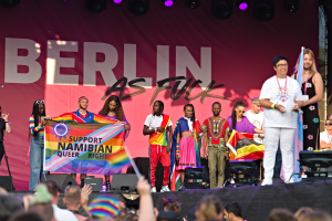 Gruppe von Menschen auf einer Bühne mit einem Banner, auf dem "Berlin as Fuck" steht, mit Lautsprechern, Mikrofonen und einer Menge unten, die Hände hebt.