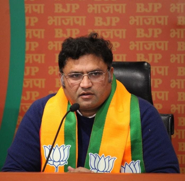 BJP-Führer Sanjay Kumar Reddy sitzt auf einem Stuhl, trägt eine Brille und einen Schal, mit einem Mikrofon auf dem Tisch vor ihm und einer Fahne mit Text im Hintergrund, während er eine Pressekonferenz in Neu Delhi abhält.