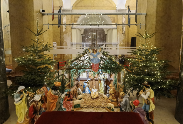 Eine Krippe in einer Kirche mit einem roten Tuch auf einem zentralen Tisch, flankiert von zwei beleuchteten Weihnachtsbäumen und einer Decke, die über einer eingezäunten Wand hängt.