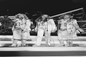 Drei Frauen singen in Mikrofone auf der Bühne mit Musikern, die hinter ihnen spielen, in Schwarz-Weiß.