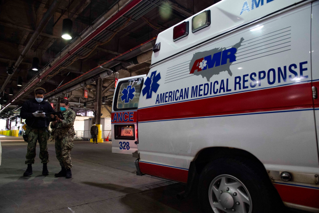 Ein Ambulanzfahrzeug der American Medical Response (AMR) steht in einem Gebäude geparkt, mit zwei maskierten Personen davor, einem Fahrzeug links daneben und mehreren Personen, Lampen, Rohren und anderen Gegenständen im Hintergrund.