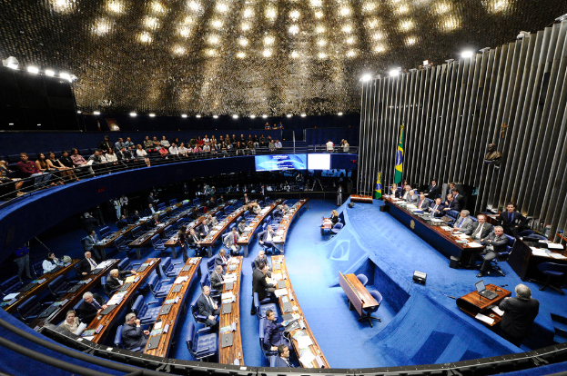 Eine Konferenzbühne mit sitzenden Personen an einem Tisch mit Papieren und Gegenständen, flankiert von Flaggen, einer Skulptur im Hintergrund und einem Publikum, das an Tischen sitzt und hinter einem Geländer mit Bildschirmen und Deckenleuchten Platz genommen hat.