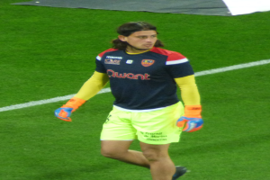 Ein Mann in einem blauen T-Shirt und gelben Shorts läuft auf einem Fußballfeld, die Arme ausgestreckt, mit einem entschlossenen Gesichtsausdruck und einem Tuch oben auf dem Bild.