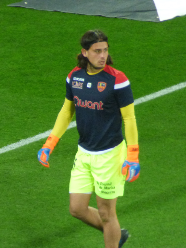 Ein Mann in einem blauen T-Shirt und gelben Shorts läuft auf einem Fußballfeld, die Arme ausgestreckt, mit einem entschlossenen Gesichtsausdruck und einem Tuch oben auf dem Bild.