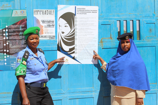 Zwei Frauen stehen nebeneinander vor einer blauen Tür, die mit Plakaten bedeckt ist, eine trägt ein blaues Hemd, schwarze Hosen und eine grüne Mütze, die andere hält ein Papier in der Hand.