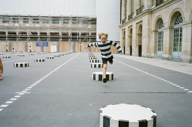 Ein Spielplatz mit vier Kindern, eines laufend auf schwarzen und weißen Betonstufen, umgeben von einem Schulgebäude.