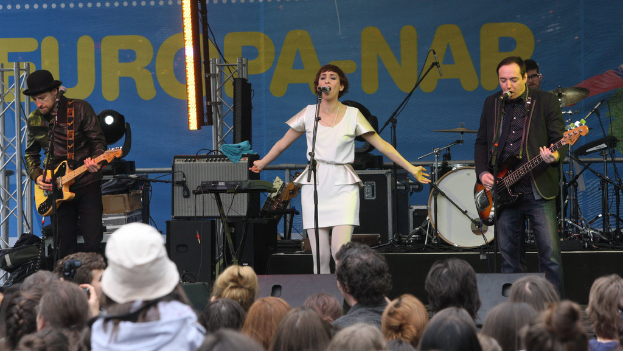 Eine Bühne mit vier Musikern: Eine singende Frau in weißem Kleid in der Mitte, zwei Gitarristen links und rechts und eine Menschenmenge und Lautsprecher im Hintergrund.