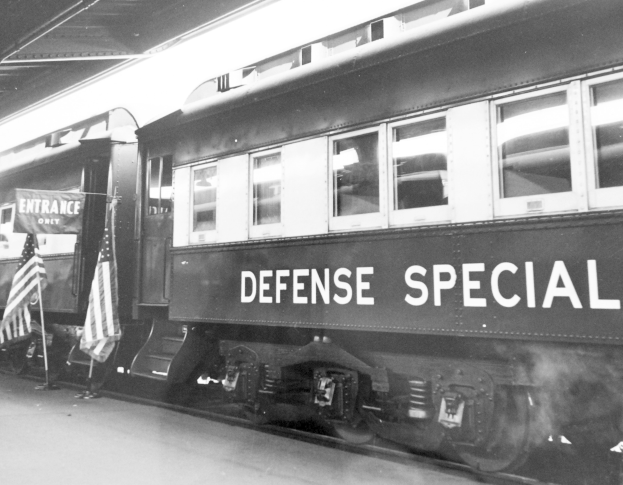 Schwarz-weißes Foto eines Zuges mit der Aufschrift "Defense Special" auf den Schienen mit zwei Fahnen links und einer Decke oben.