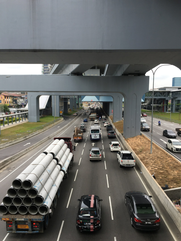 Eine stark befahrene Autobahn mit viel Verkehr unter einer Brücke, umgeben von Gras, Bäumen, Gebäuden, Pfählen und Straßenlaternen unter einem klaren Himmel.
