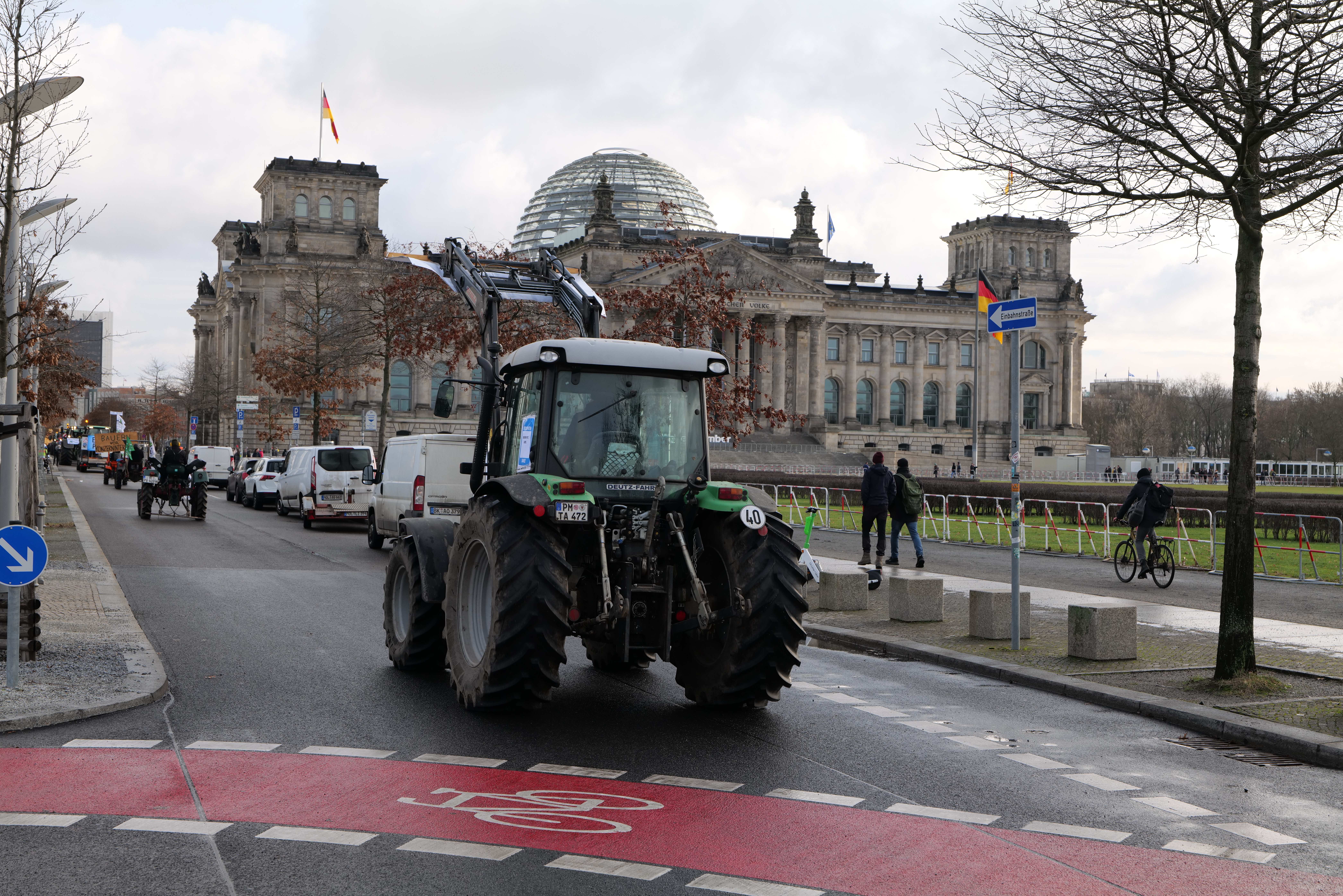 Ein Traktor fährt eine Straße vor dem Reichstaggebäude in Berlin, Deutschland, entlang, während Menschen auf den Gehwegen gehen und Fahrräder fahren. Bäume, Laternenmasten und Schilder säumen die Straße, und Fahnen schmücken das Gebäude bei einem bewölkten Himmel.