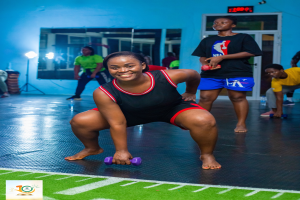 Eine Frau in einem Fitnessstudio ist in einer Kniebeugeposition und hält Hanteln, mit verschiedenen Fitnessgeräten und Logos im Hintergrund.