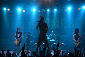 Eine Gruppe von Musikern performs auf einer hell erleuchteten Bühne mit Gitarren, einem Mikrofon und Trommeln, während die Hände des Publikums im Vordergrund an der O2 Arena zu sehen sind.