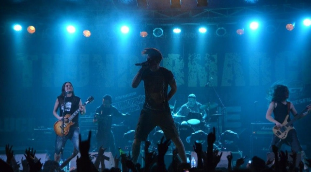 Eine Gruppe von Musikern performs auf einer hell erleuchteten Bühne mit Gitarren, einem Mikrofon und Trommeln, während die Hände des Publikums im Vordergrund an der O2 Arena zu sehen sind.