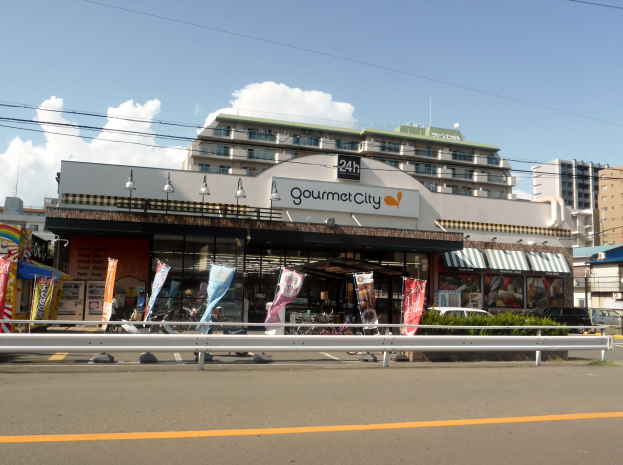 Eine belebte Stadtstraße mit Gebäuden, Straßenmöblierung, Fahrzeugen, Grünflächen und einem Restaurant mit Essensauslage.