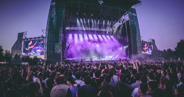 Eine große Menschenmenge versammelte sich auf einem Musikfestival, mit einer Bühne in der Mitte, auf der Musiker Instrumente spielten, beleuchtet von Lichtern und umgeben von Bildschirmen, vor einer Kulisse aus einem Gebäude, Bäumen und einem klaren blauen Himmel.