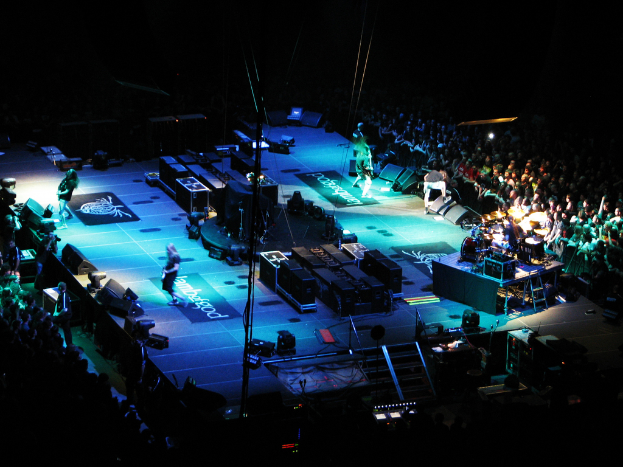 Eine Gruppe von Menschen, die auf einer Bühne mit Lautsprechern, Scheinwerfern und Mikrofonen performs, mit einer großen Menge auf beiden Seiten vor einem dunklen Hintergrund.