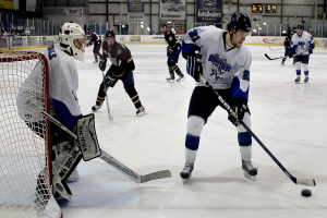 Eine Gruppe junger Männer spielt Eishockey auf einem Eis, trägt Helme und hält Hockey-Schläger, mit einem Torpfosten links und Bannern im Hintergrund.