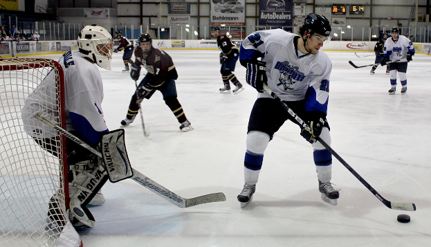 Eine Gruppe junger Männer spielt Eishockey auf einem Eis, trägt Helme und hält Hockey-Schläger, mit einem Torpfosten links und Bannern im Hintergrund.