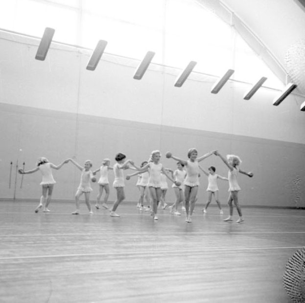 Schwarzes Bild von jungen Mädchen, die Hand in Hand tanzen und in einem Studio mit einem Ball auf der rechten Seite und Lampen an der Decke.