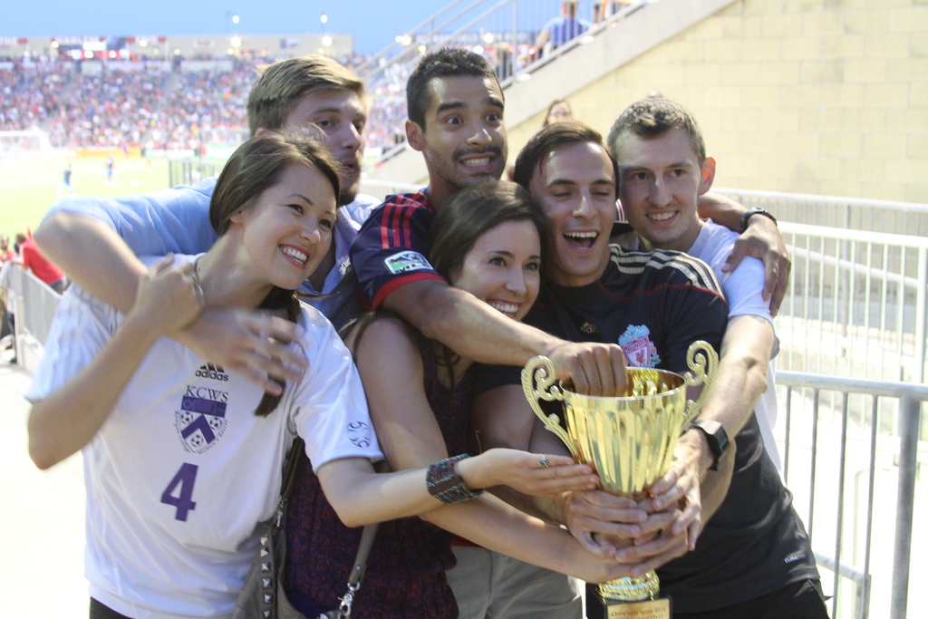 Sechs Menschen, darunter zwei Frauen und vier Männer, halten einen Pokal und lächeln, während sie für ein Foto vor einem Eisengeländer posieren, mit einer Wand und einem Geländer oben rechts, Menschen und Lichter im Hintergrund, möglicherweise in einem Stadion.