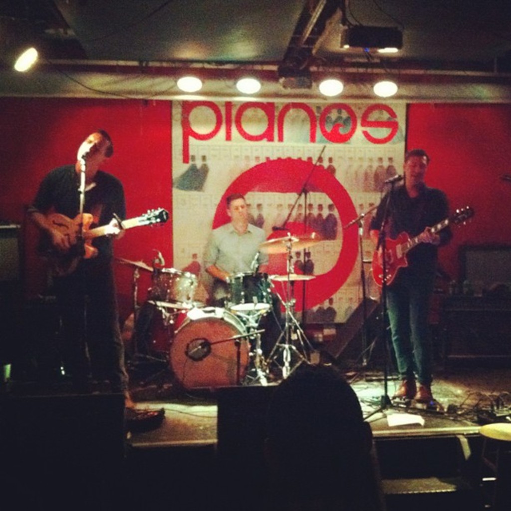 Drei Männer auf der Bühne: Einer spielt am Drumset in der Mitte, zwei spielen Gitarre und singen auf der linken und rechten Seite, mit einem Banner an der Wand und Lampen an der Decke.