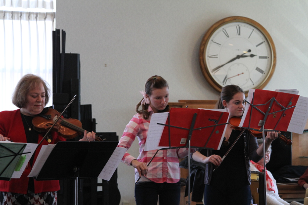 Drei Frauen auf der Bühne mit Geigen stehend und auf Musikständern spielend, während ein sitzendes Publikum zuhört; eine Uhr hängt an der weißen Wand hinter ihnen, und ein weißer Vorhang ist links sichtbar.