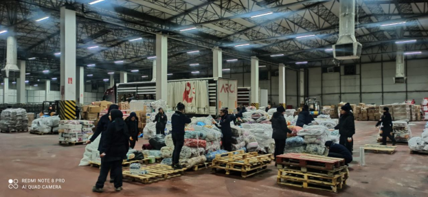 Eine Gruppe von Menschen steht vor einem großen Lagerhaus, das mit Säcken voller Lebensmittel, Holzpaletten, Containern, Polen, einem Schild, einer Wand, einem Dach mit Deckenleuchten und Metallstangen gefüllt ist, und ein Wasserzeichen auf der linken Seite zu sehen ist, das den Anschein eines Lebensmittelverteilungszentrums in Dubai erweckt.