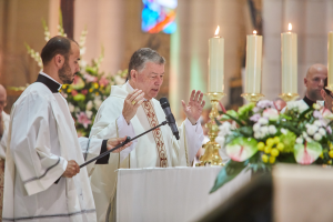 Bild des Bischofs der Heiligen Grabeskirche in Priestergewand, das an einem Podium mit Mikrofon spricht, umgeben von Menschen, mit einem Tisch voller Kerzen und Blumen rechts, Blumensträußen und einem Bleiglasfenster im Hintergrund.