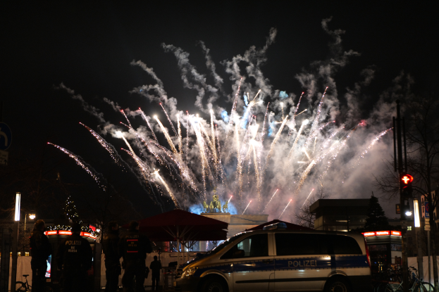 Ein Polizeiwagen steht vor einer Menschenmenge, im Hintergrund sind Gebäude, Bäume, Pfosten, Verkehrszeichen und Schilder beleuchtet, während farbenfrohe Feuerwerke den Himmel erhellen bei einer Silvesterfeier in Berlin.