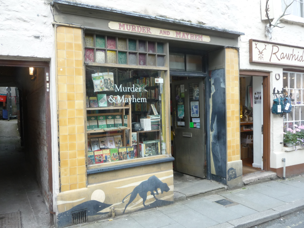 Ein Geschäft mit dem Namen 'Mord und Totschlag' mit Regalen voller Bücher, verschiedener Gegenstände, Texttafeln und Pflanzen mit Blumen, einem Baum und einer Straße im Vordergrund.