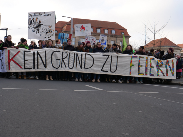 Eine Gruppe von Menschen steht auf der Straße und hält ein Transparent mit der Aufschrift "Kein Grund zu Feiern" im Protest, mit Straßenlaternen, Bäumen, Gebäuden mit Fenstern und einem klaren blauen Himmel im Hintergrund.