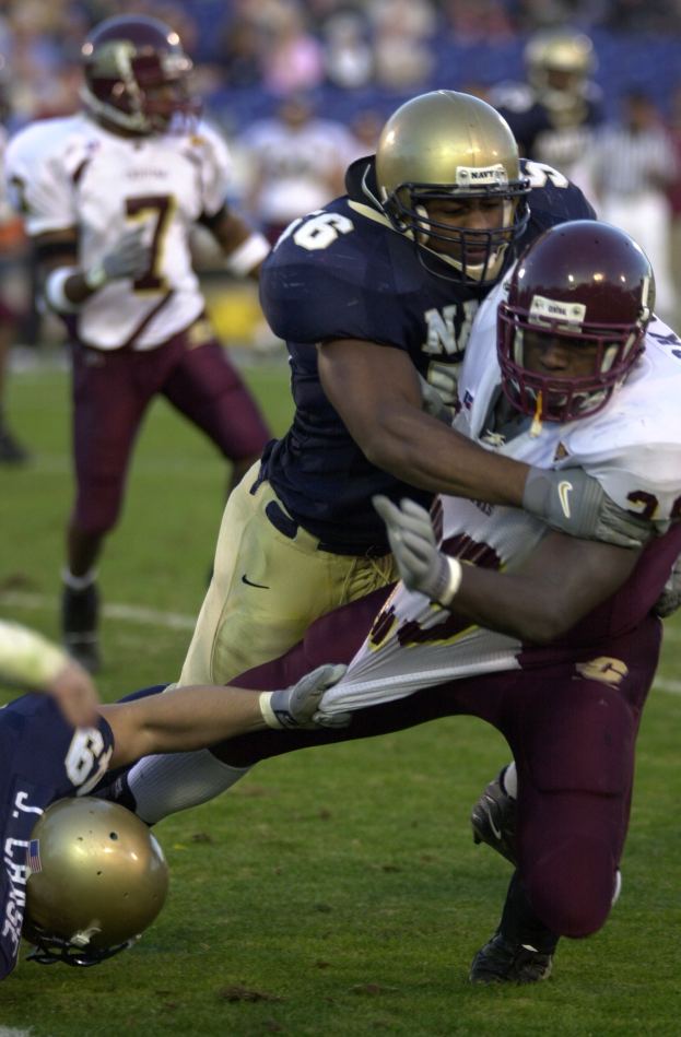 Zwei Fussballspieler in Helmen und Uniformen räumen sich gegenseitig auf dem Feld, während Teamkollegen und Zuschauer im Hintergrund zusehen.