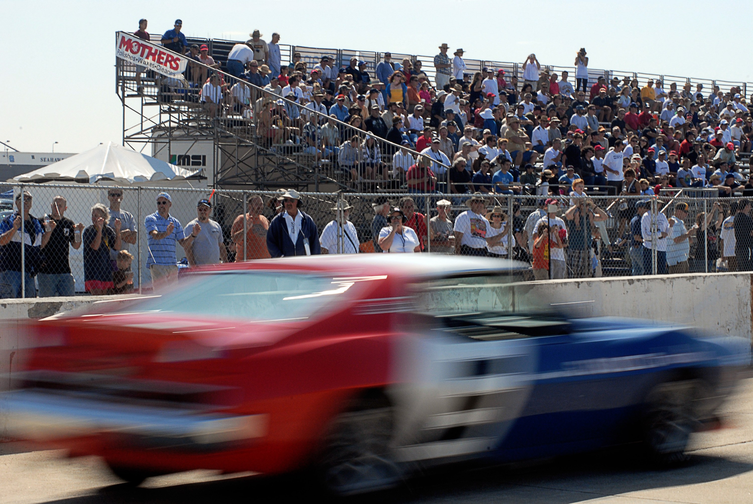 Ein rotes und blaues Auto, das auf einer Rennstrecke vor einer Zuschauermenge fährt, mit Maschendrahtzaun, Treppen, Bannern, Zelten und Laternen im Hintergrund.