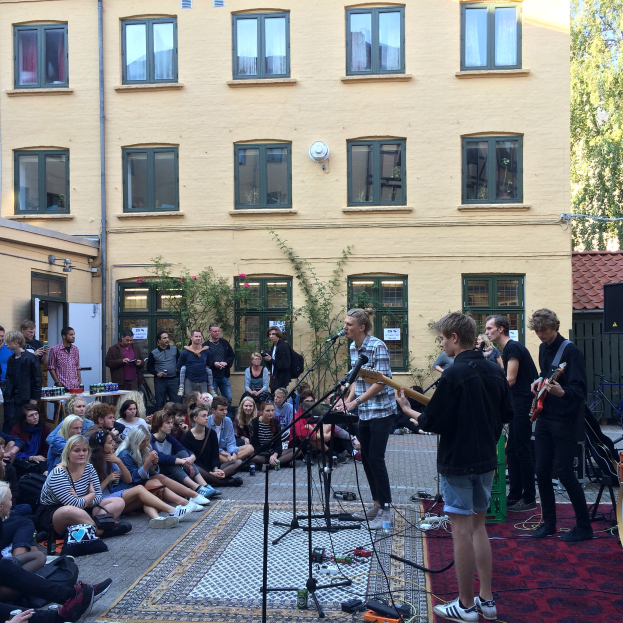 Eine Gruppe von Menschen sitzt auf dem Boden und spielt mit Musikinstrumenten, Mics und verstreuten Gegenständen vor einem Gebäude in Berlin.