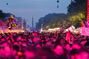 Eine große Menschenmenge bei einem nächtlichen Musikfestival, beleuchtet von bunten Lichtern und Ballons, mit Zelten, Bannern, Fahnen und anderen Gegenständen, die herumliegen, Bäume, ein Turm und ein sternenübersäter Himmel im Hintergrund.