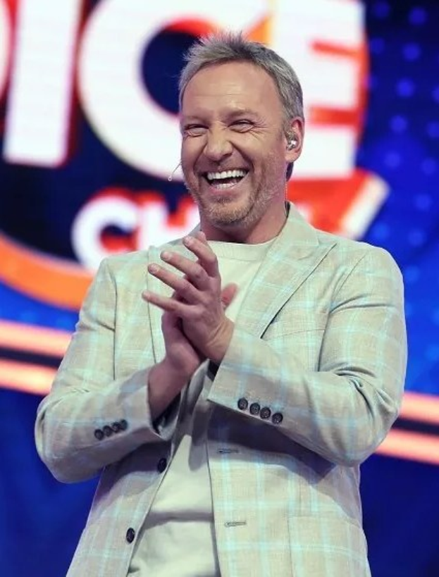Ein lachender Mann in einem Anzug, der auf einer Bühne klatscht, mit dem 'The Voice'-Logo im Hintergrund.