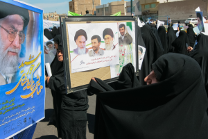 Eine Gruppe von Frauen in schwarzen Abayas hält Plakate mit Bildern von Ayatollah Ali Khamenei hoch, mit Gebäuden, Pfählen, Fahrzeugen und einem klaren blauen Himmel im Hintergrund.