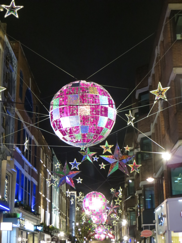 Eine Stadtstra├če bei Nacht mit zahlreichen Lichtern und Deko, mit einem gro├čen Diskokugel an einem Geb├Ąude h├Ąngend, die eine festliche Atmosph├Ąre schafft.