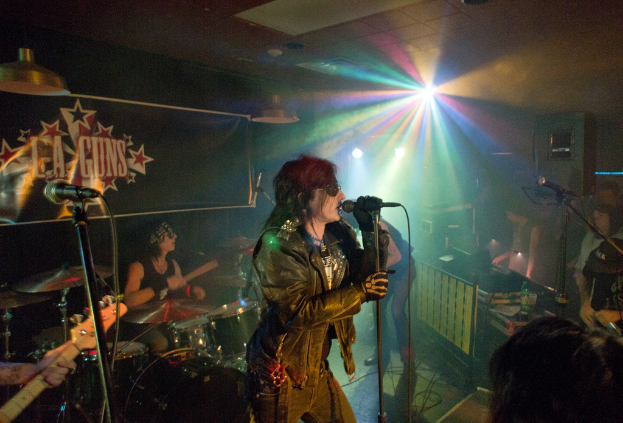 Eine Frau in einer schwarzen Jacke singt auf der Bühne in ein Mikrofon, während ein Mann mit Sonnenbrille und Stirnbinde ein Instrument spielt und ein Plakat mit "LA Guns" im Hintergrund zu sehen ist.