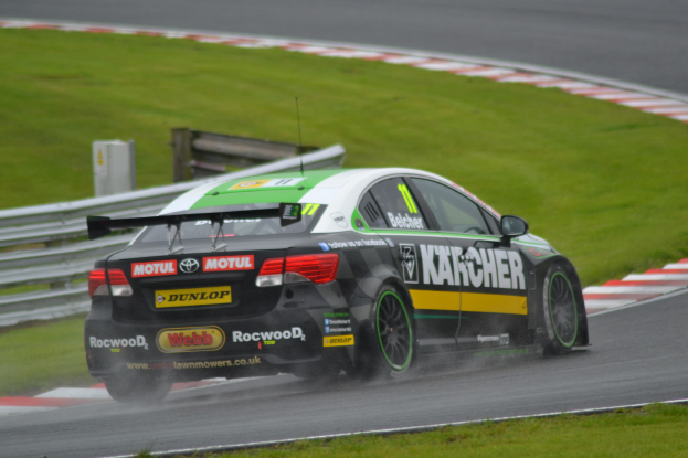 Ein Auto mit Text an der Seite fährt auf einer regennassen Rennstrecke, die von Gras gesäumt ist, mit einem Geländer und einer hölzernen Konstruktion im Hintergrund.