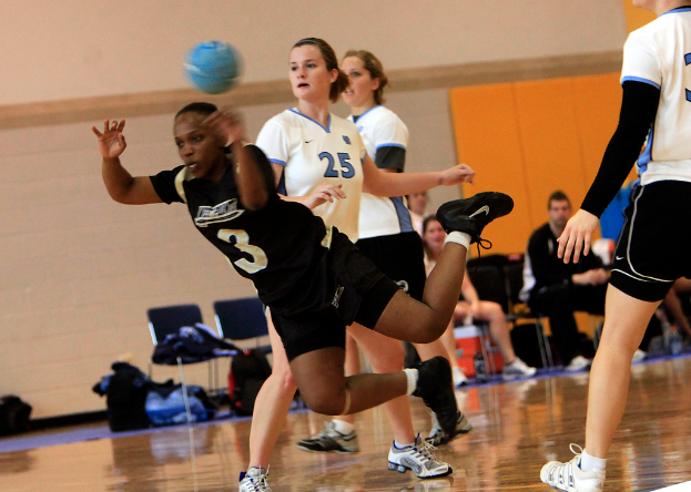 Mehrere Mädchen spielen mit einem blauen Ball auf einem Indoor-Court, während andere Menschen auf Stühlen in der Nähe sitzen, mit Gepäck und einer Wand im Hintergrund.