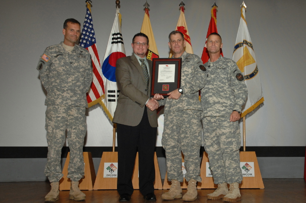 Vier Männer auf einer Bühne: drei in Armeeuniformen und einer in Blazer, Krawatte und Hemd, die Hände schütteln und einen Preis überreichen, mit sechs Fahnen auf Stangen und einer weißen Wand im Hintergrund.